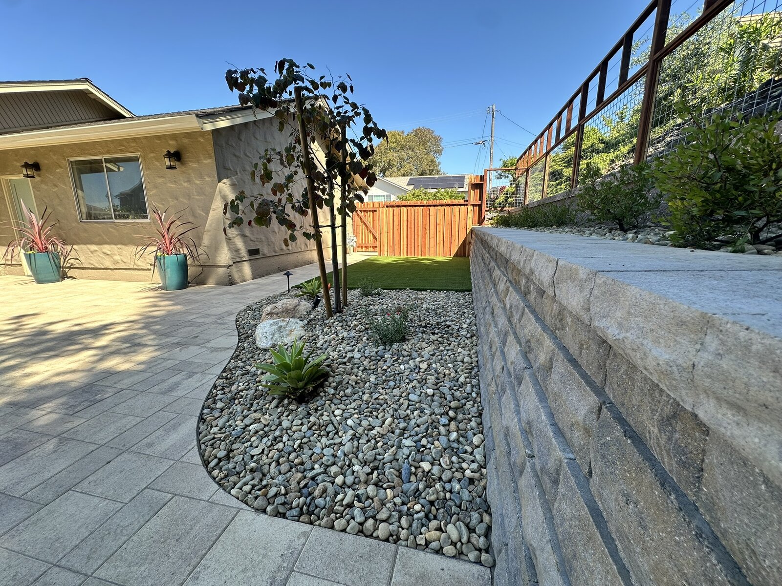 Planting bed completed — retaining wall with river rock, young tree, and paver patio